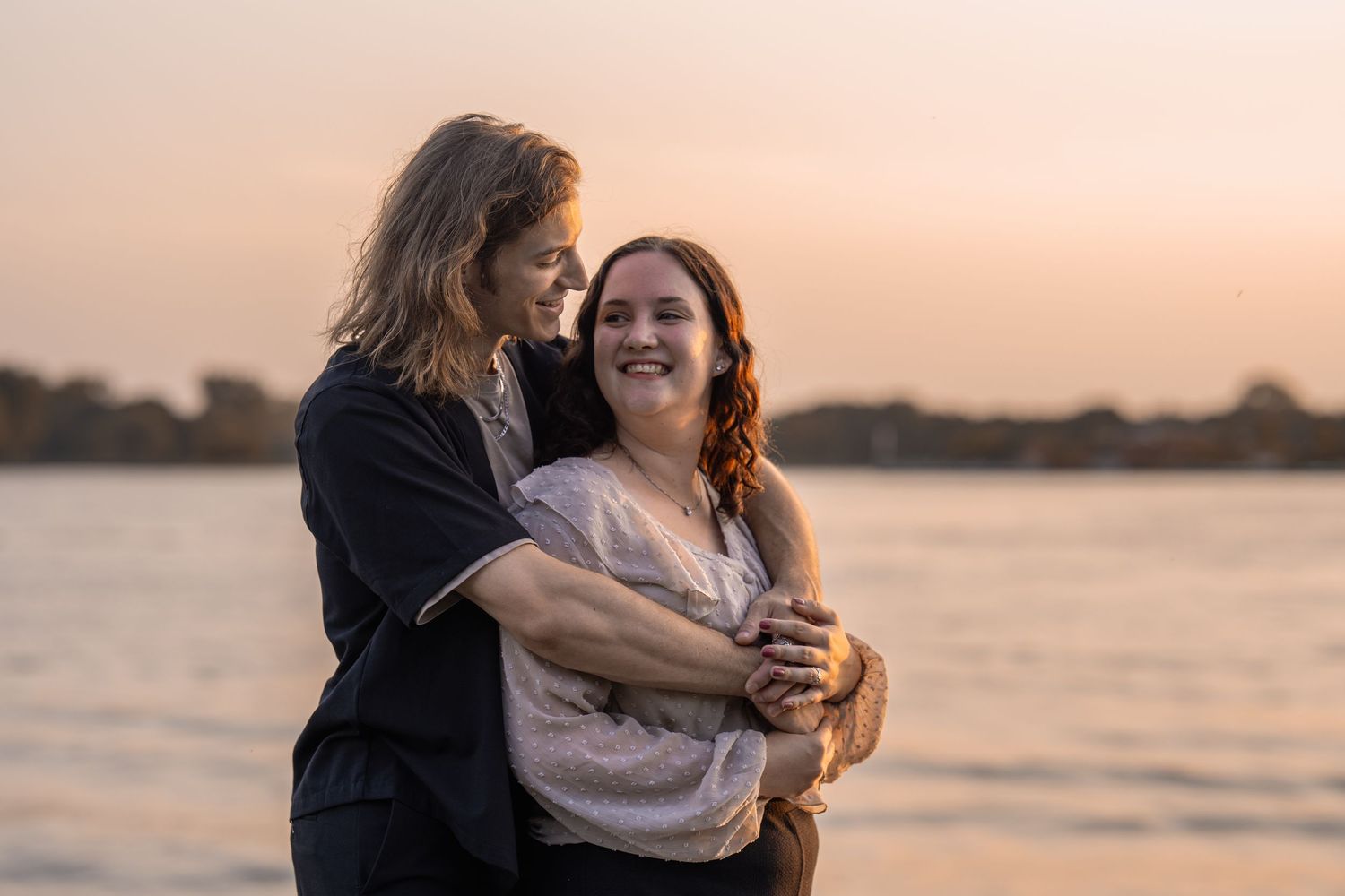Two women embrace and smile at sunset by a lake, sharing a joyful moment in warm golden light.