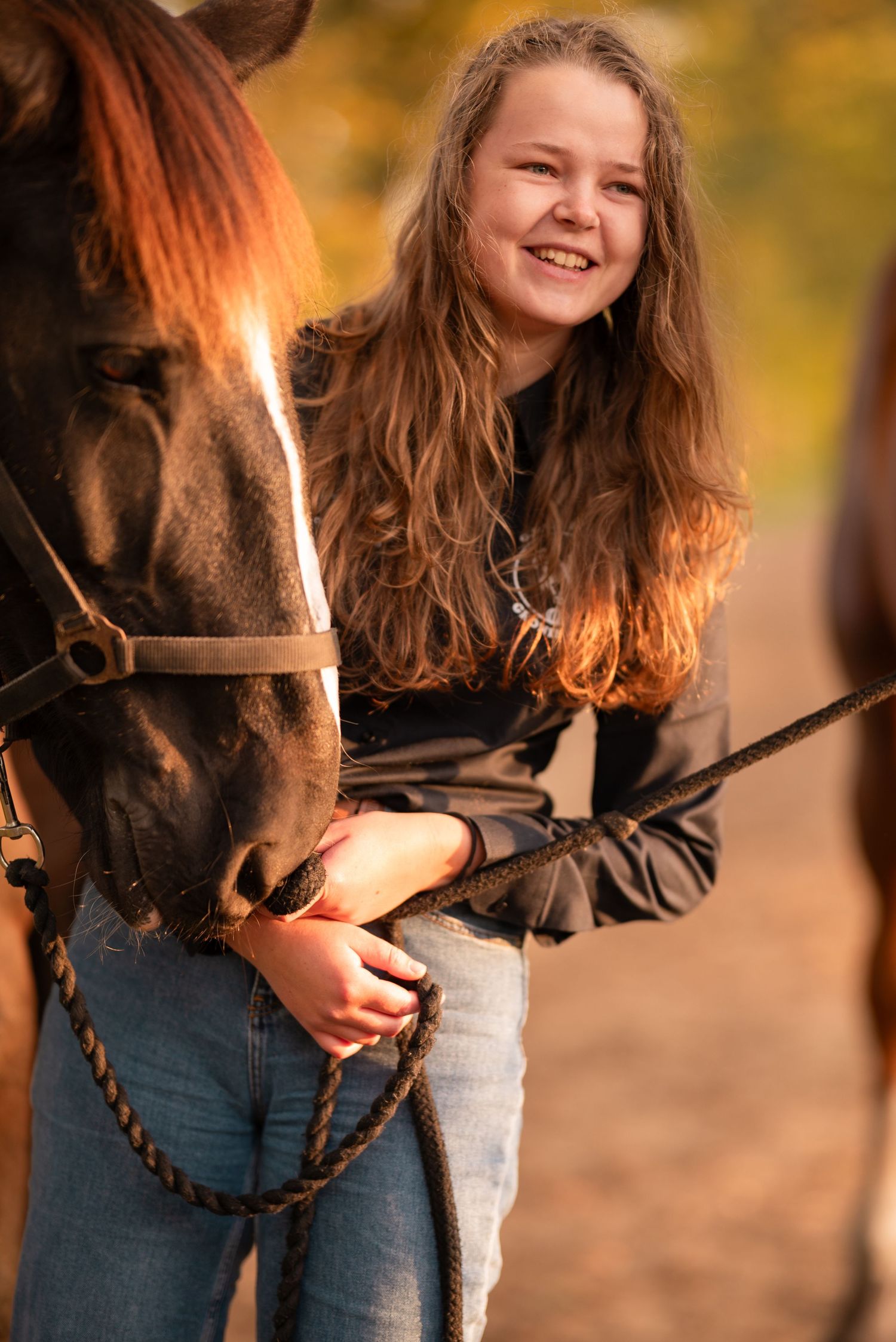 Smiling teenage girl with long brown hair holding the reins of a dark horse outdoors in warm autumn light.
