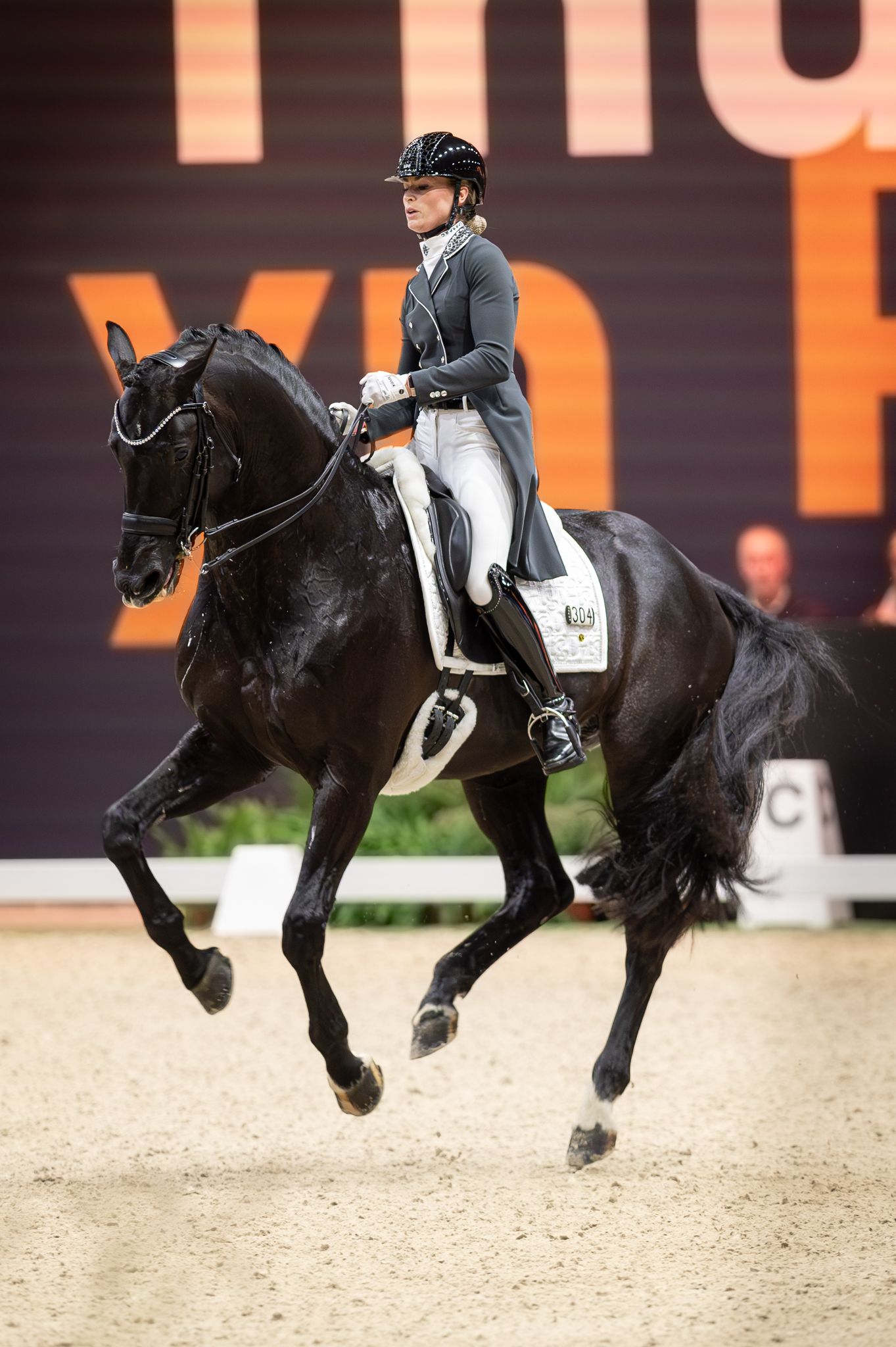 A female equestrian in formal attire rides a black horse performing dressage in an indoor arena.