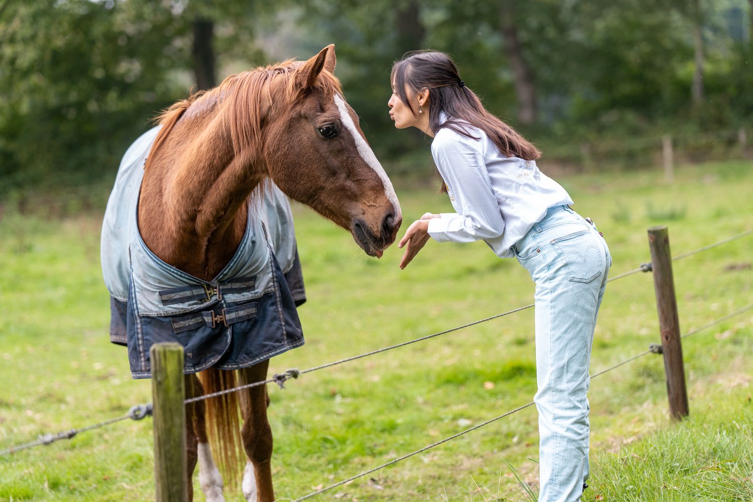 Young woman in white shirt leaning toward a chestnut horse at a fence.