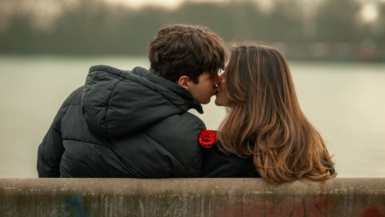 A young couple shares a romantic kiss outdoors near a lake, man wearing black jacket with red rose.