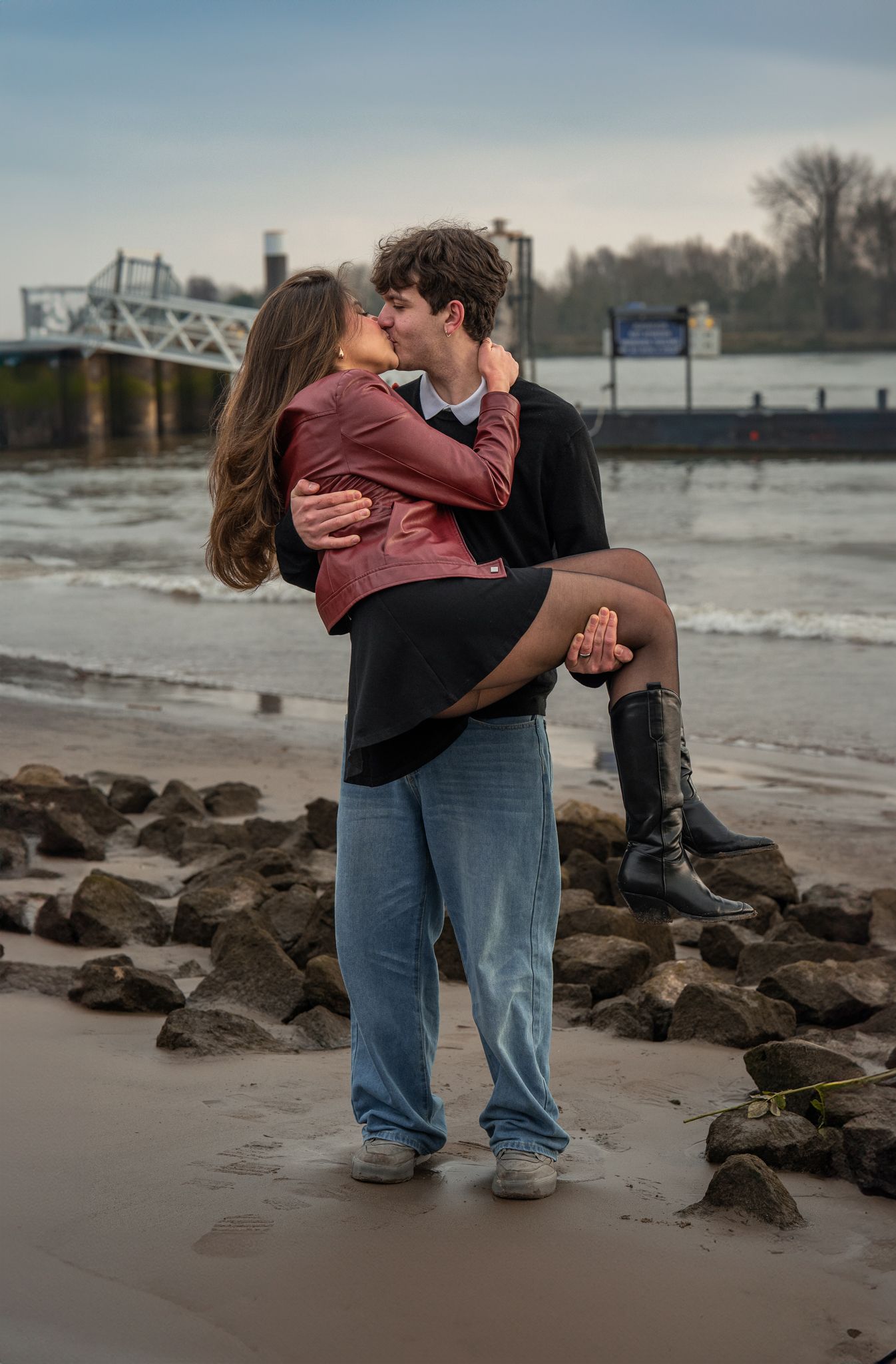 A couple shares a romantic kiss on a rocky beach, the man lifting the woman in his arms near the water.