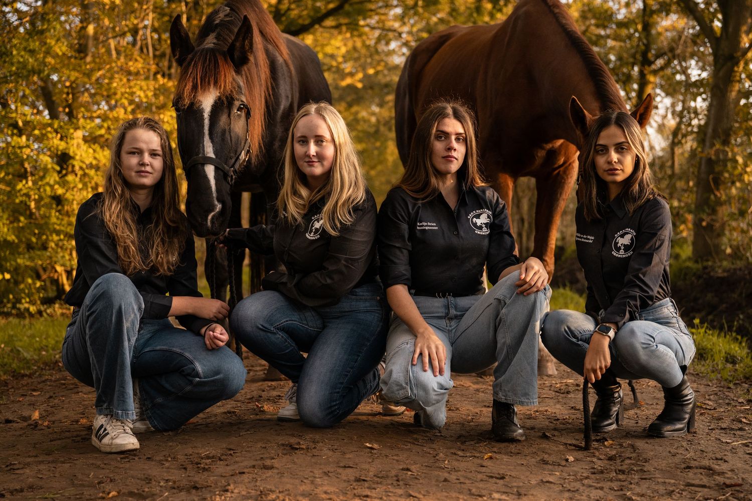 Four young women posing with two brown horses on a dirt path surrounded by autumn foliage.