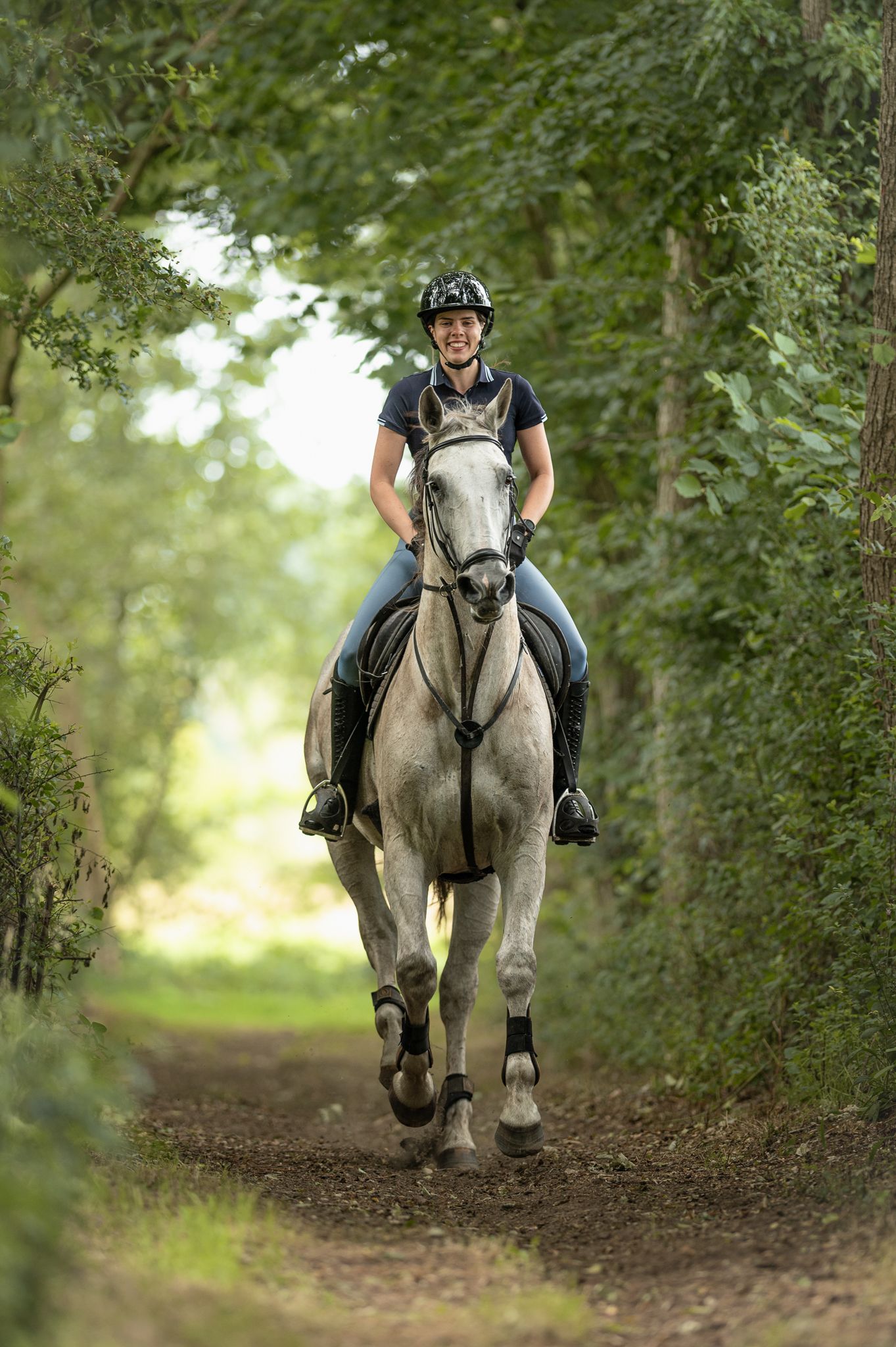 Smiling female rider on a grey horse trotting down a tree-lined forest trail.