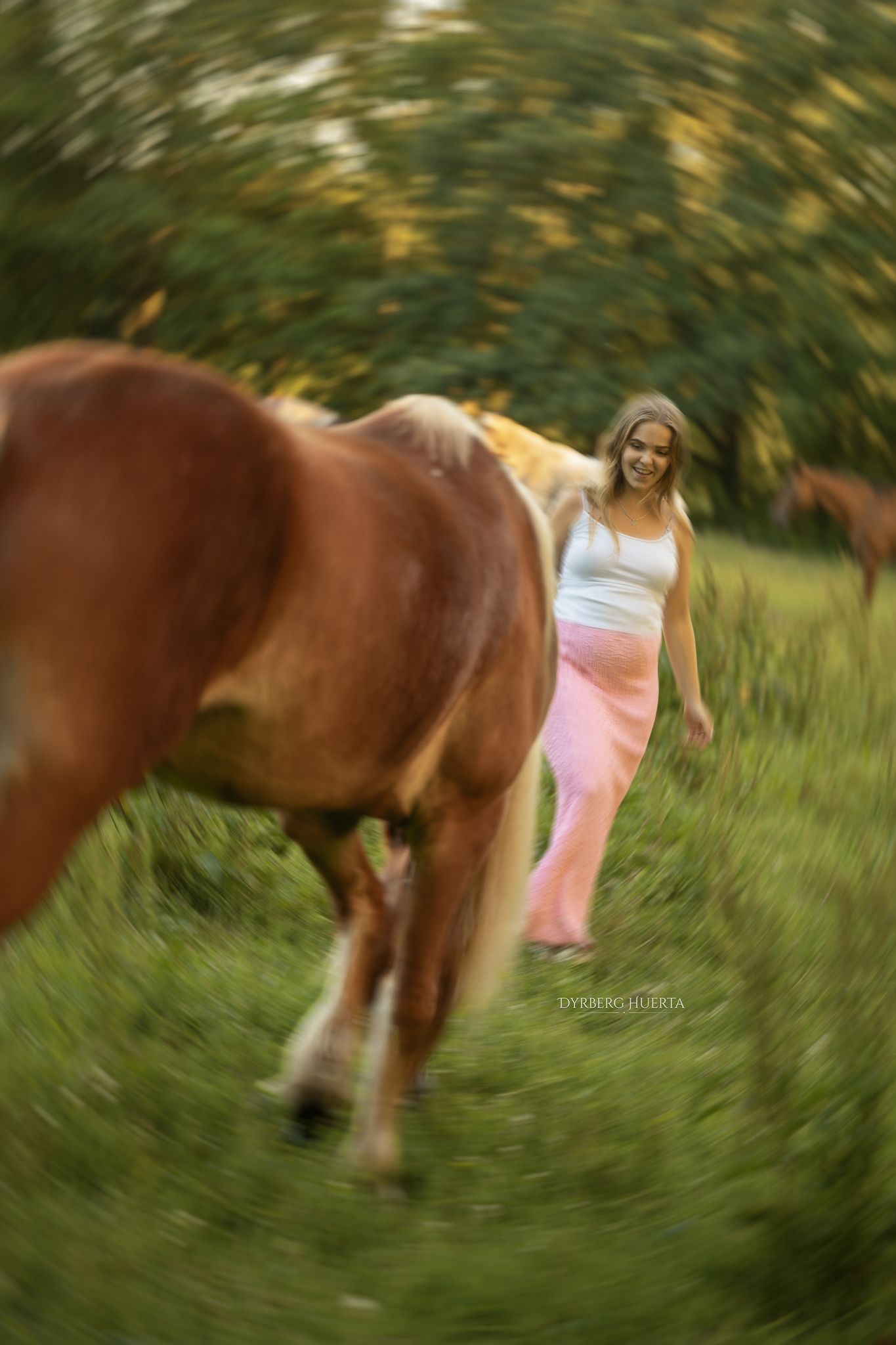 Pregnant woman in pink skirt walks alongside brown horses in a lush green field with bokeh effect.