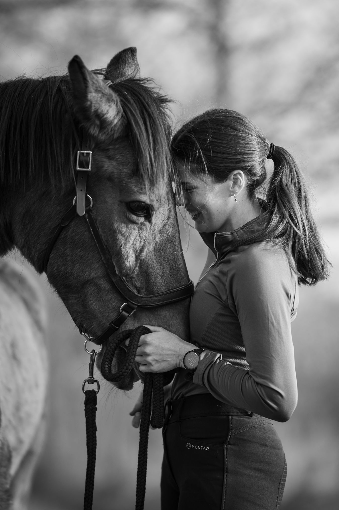 Black and white portrait of a smiling woman tenderly bonding with her horse outdoors.