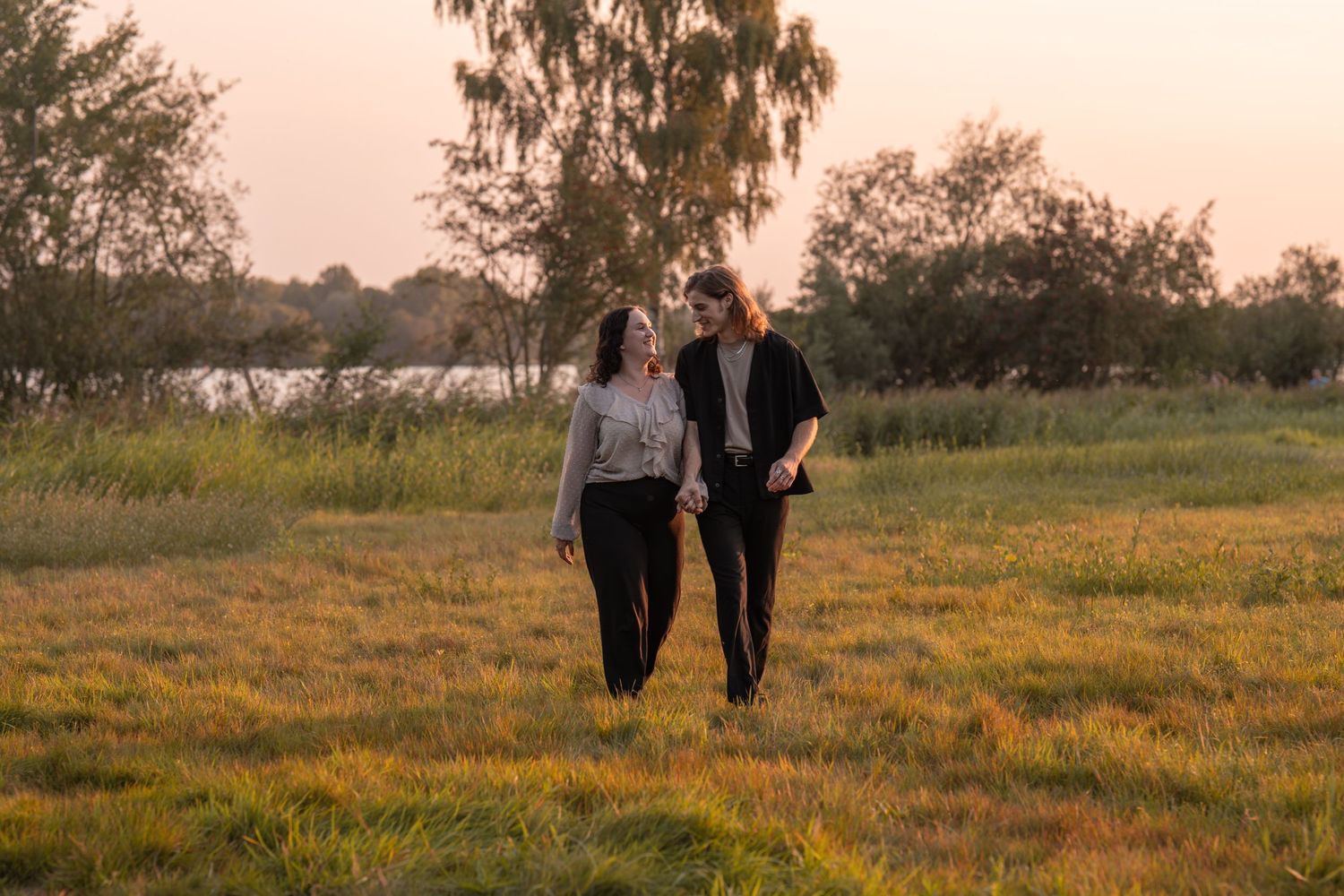 Couple walking hand in hand through a golden meadow at sunset with trees and a lake in the background.