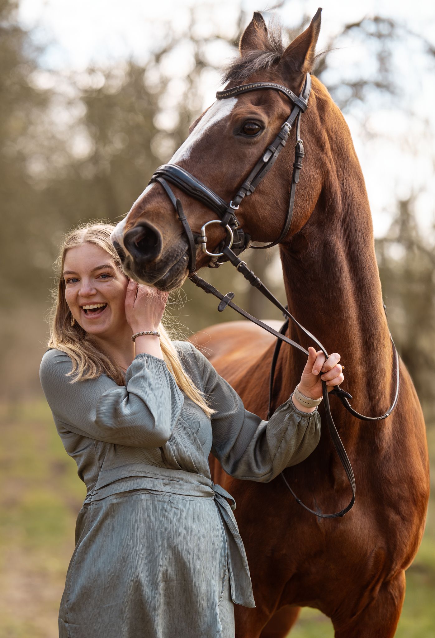 Laughing blonde woman posing with a tall chestnut horse wearing a bridle outdoors.