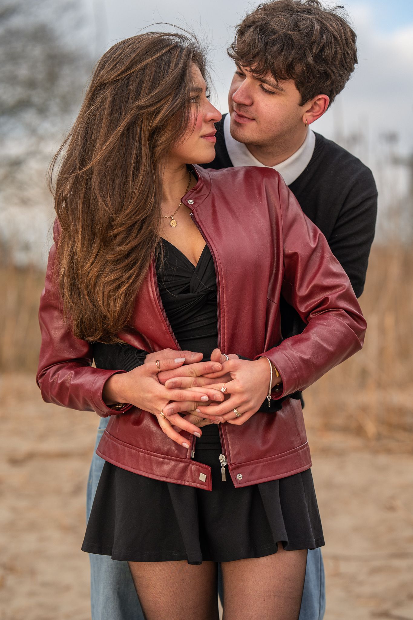 Young couple embracing outdoors, woman in red leather jacket and black dress held from behind.