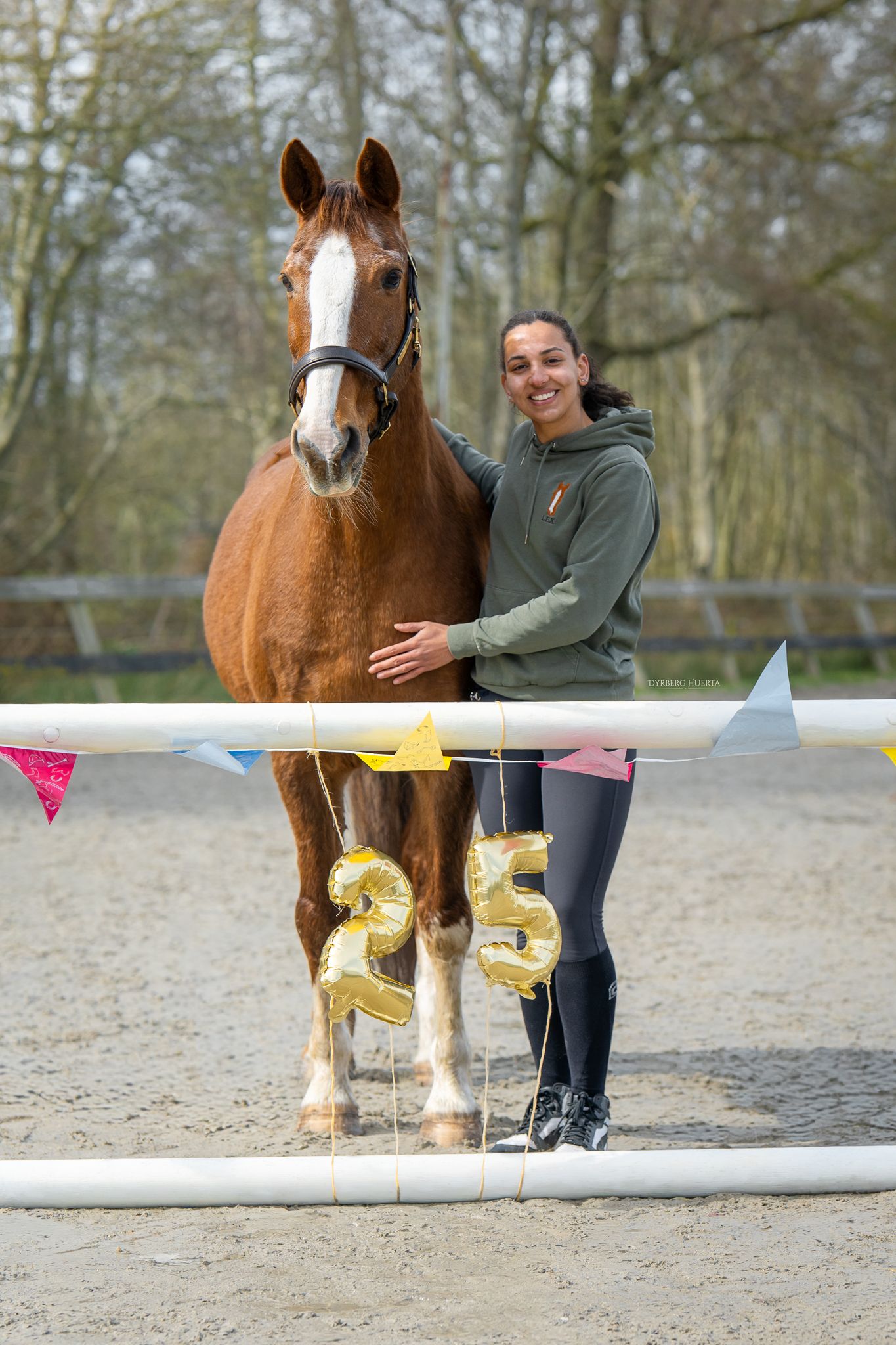 A smiling woman hugging a chestnut horse decorated with gold number 25 balloons in an arena.