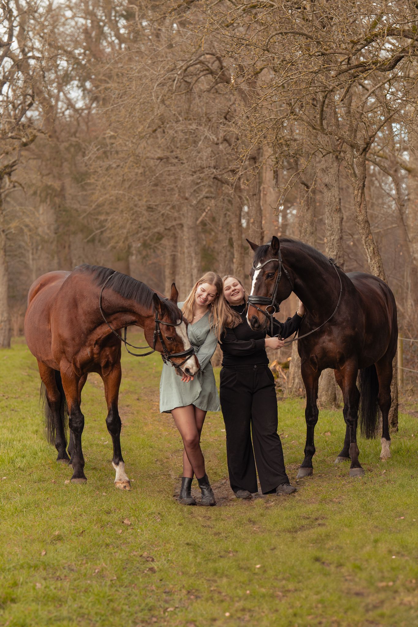Two blonde women stand smiling between two brown horses in a bare autumn forest setting.