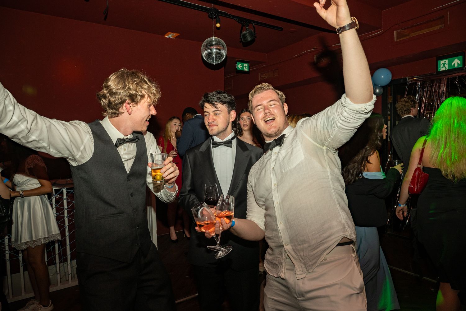 Three young men in formal attire celebrate at a party, dancing and holding drinks under disco lights.