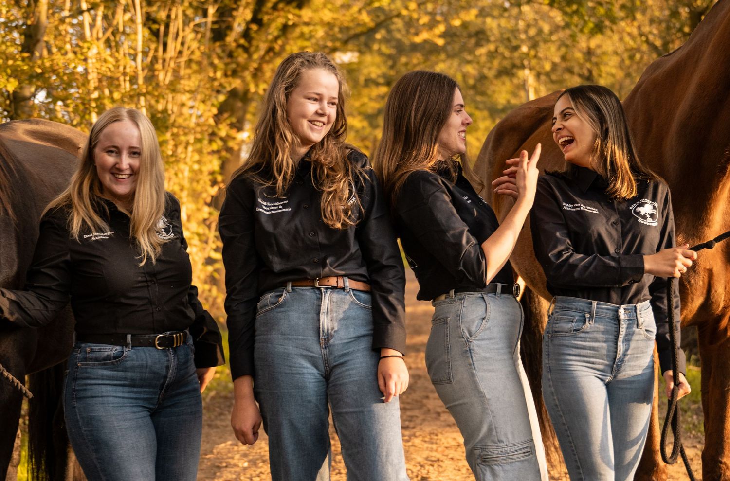 Four young women in matching black shirts laughing together outdoors with horses in autumn light.