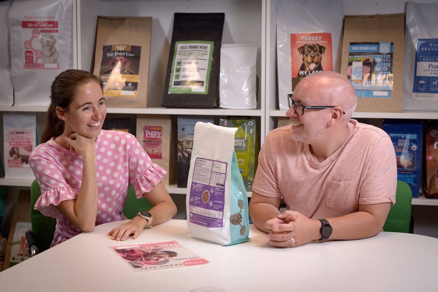Two people sit at desk with bag of dog food on table , shelves of products behind, office setting.