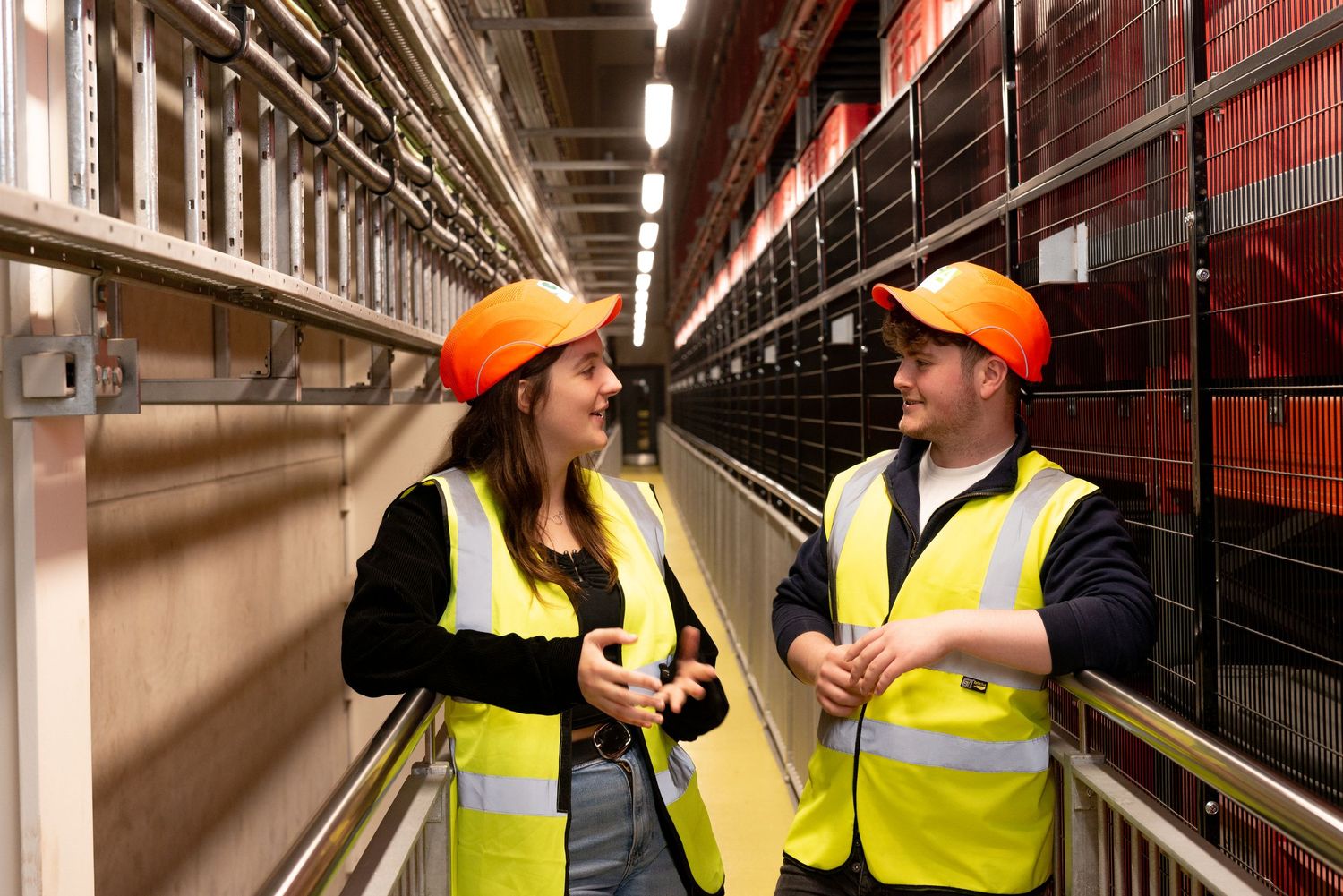 Two young workers in hi vis chat in warehouse gantry.