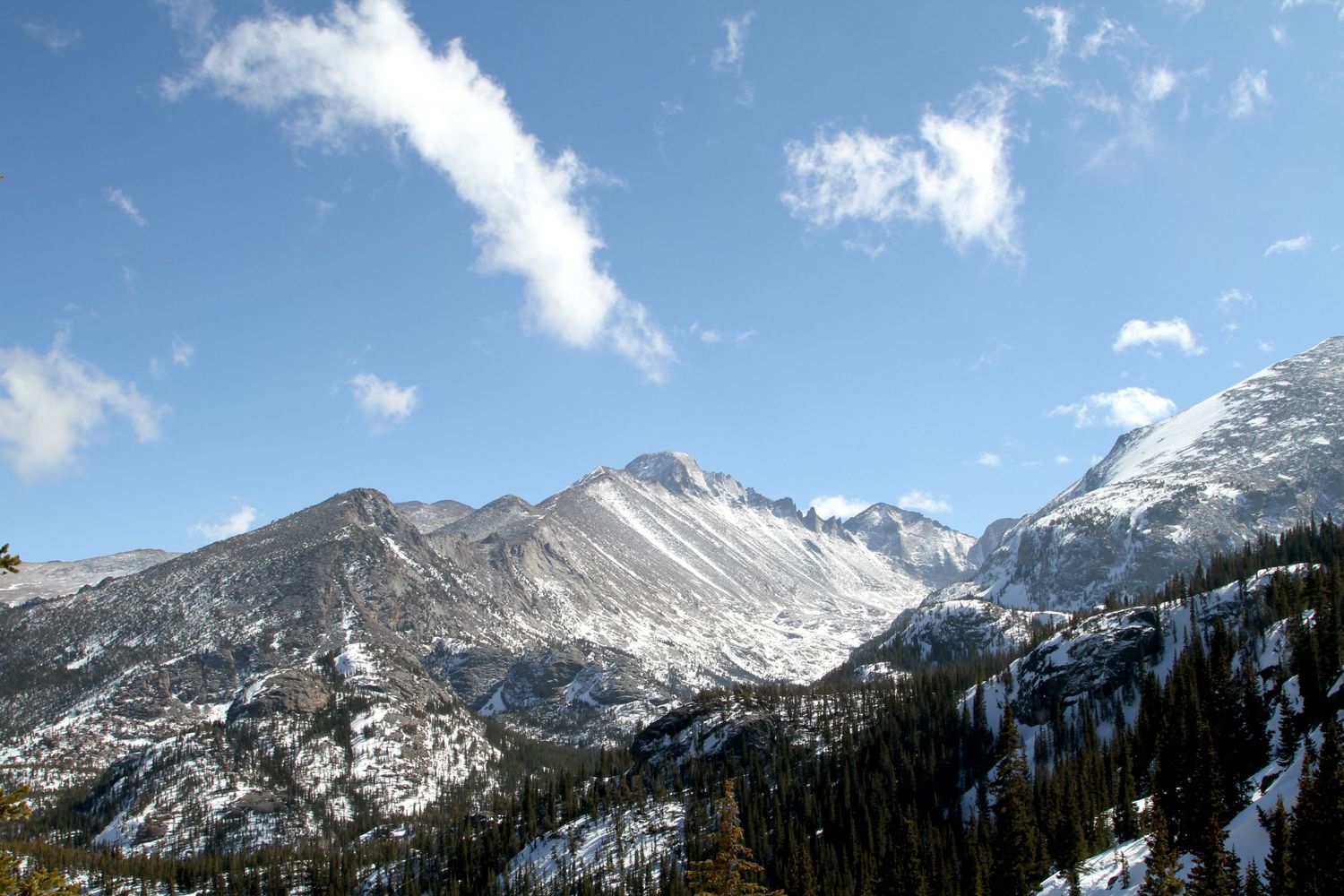 Snow-covered mountain peaks rise above evergreen forest under bright blue sky with white puffy clouds in winter landscape.