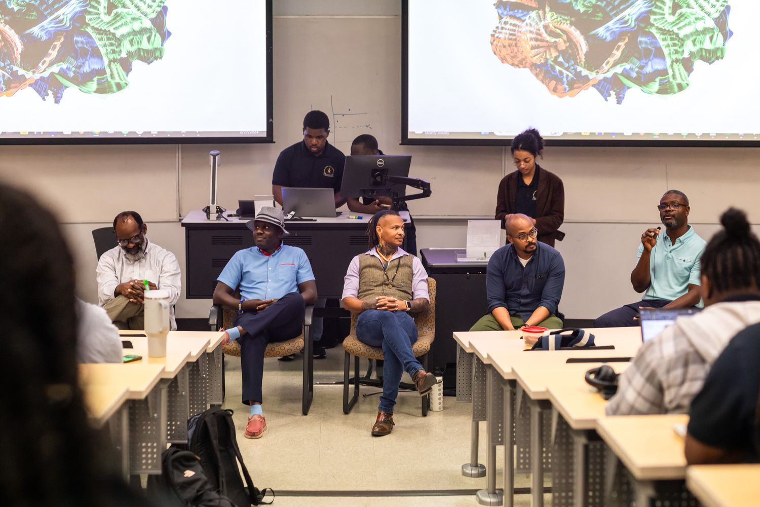 A large group meets in an auditorium-style classroom with wooden slat walls and presentation screens.