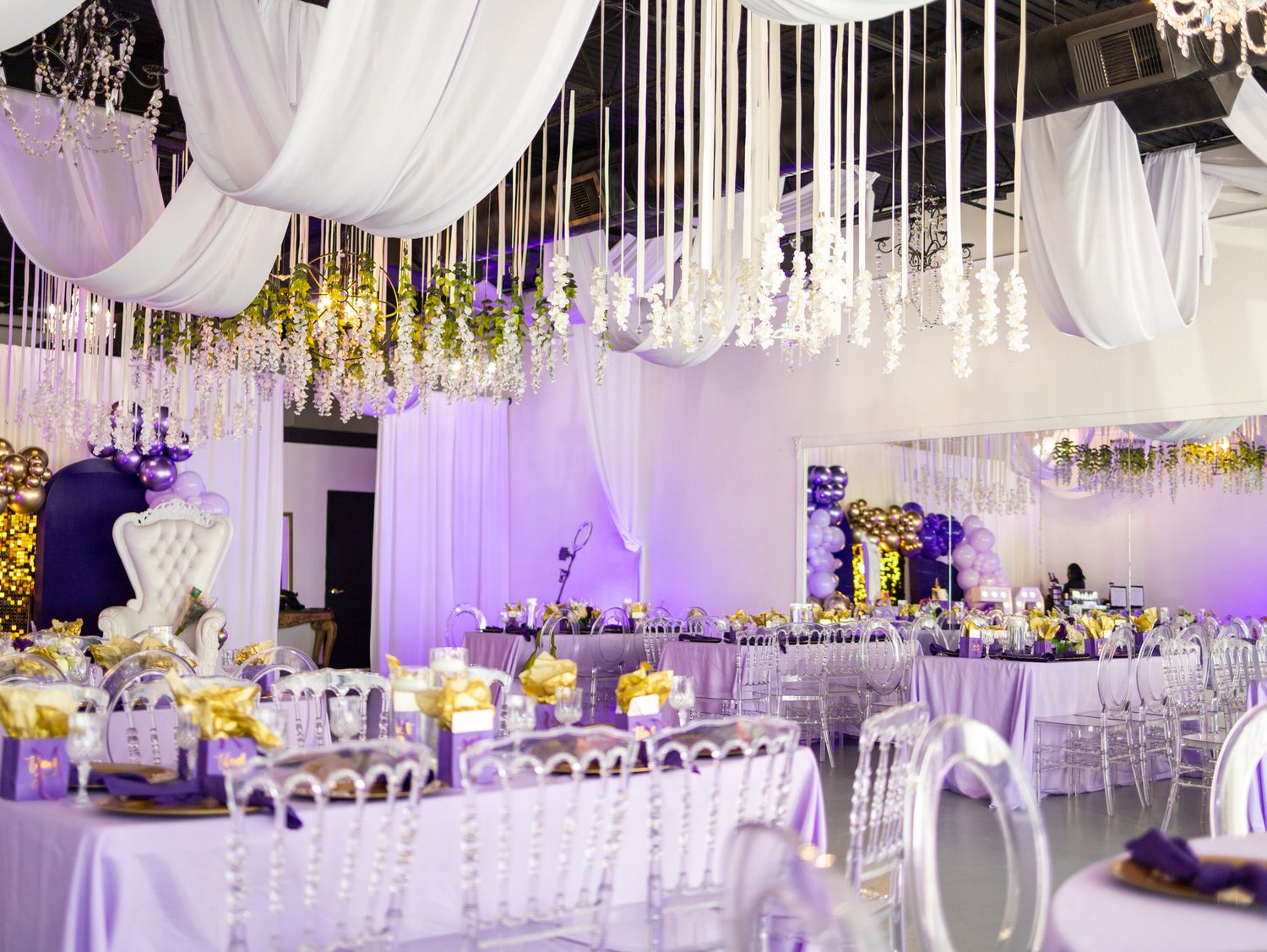 Elegant wedding reception venue with purple lighting, white drapes, and crystal chandeliers hanging from the ceiling.