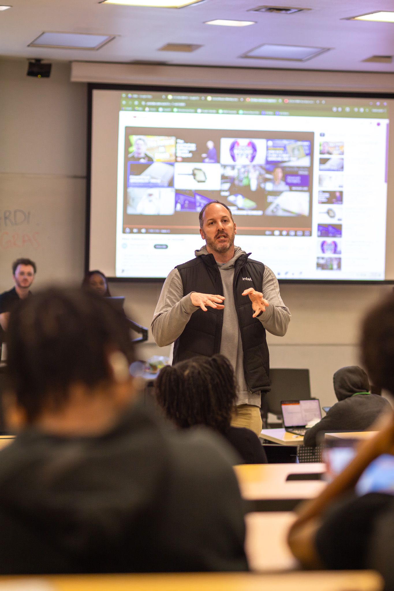 Speaker presents to an audience with projected screens visible in the background.