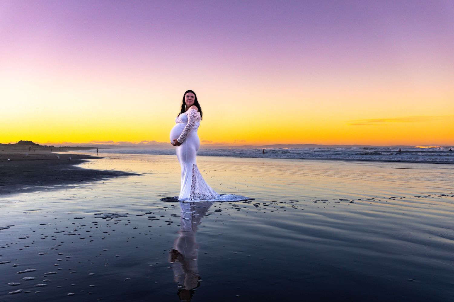Pregnant woman in white lace gown standing on beach at sunset with vibrant purple and orange sky reflected on wet sand.