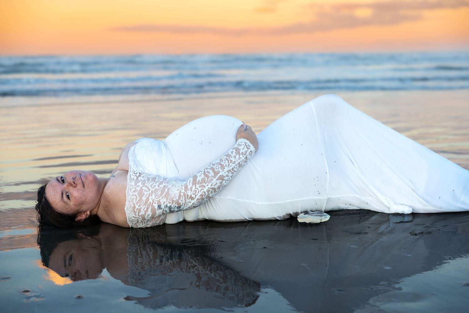 Bride in white lace gown lying on wet beach sand at sunset with ocean waves and colorful sky reflection.