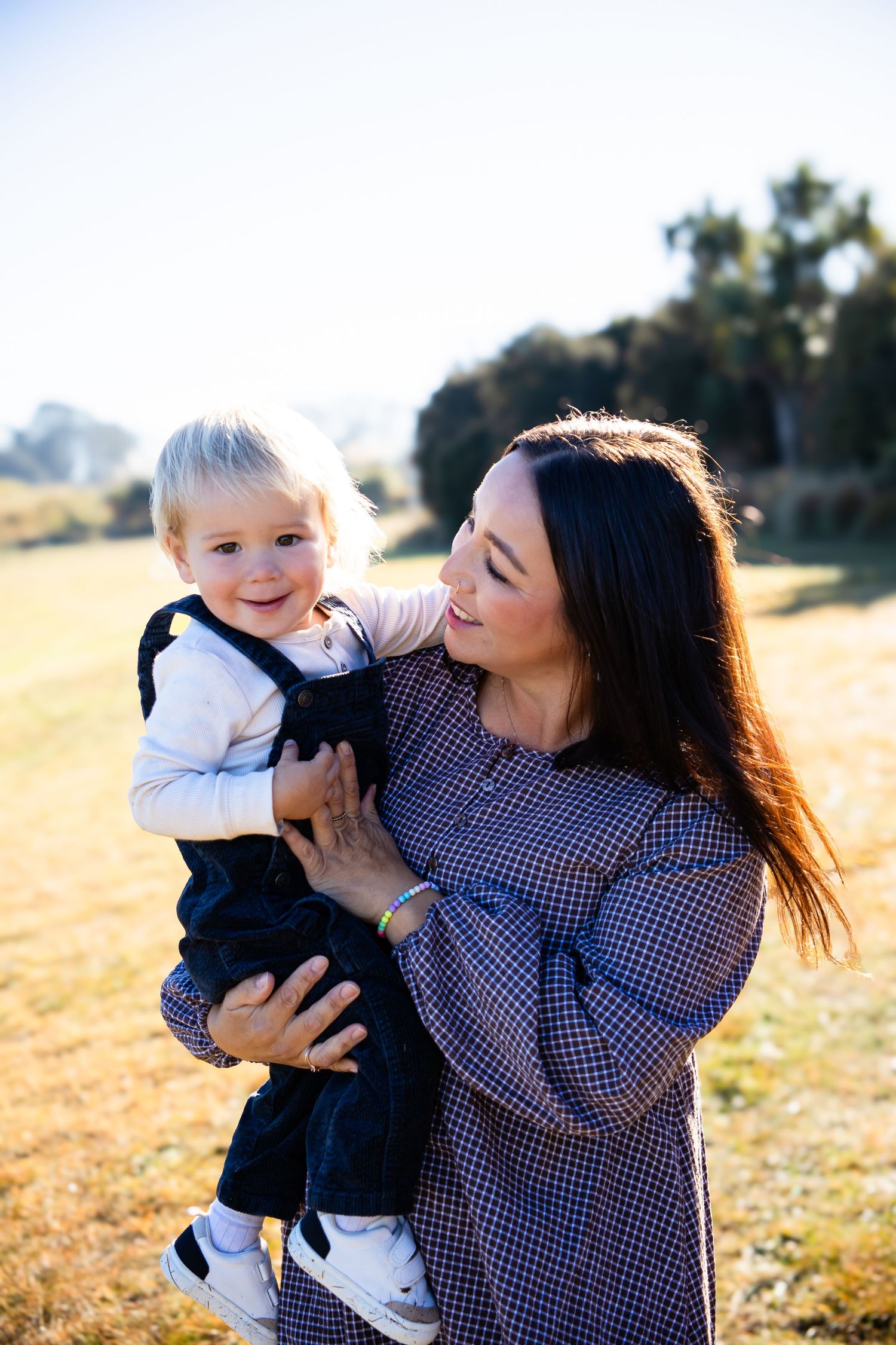 Mother holding smiling toddler boy in black overalls outdoors in sunny field.