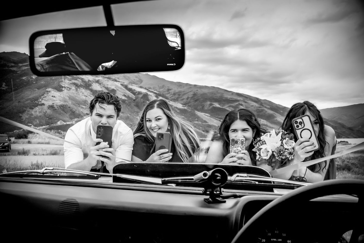 Black and white photo of four friends looking at phones in a convertible car with scenic mountains in the background.