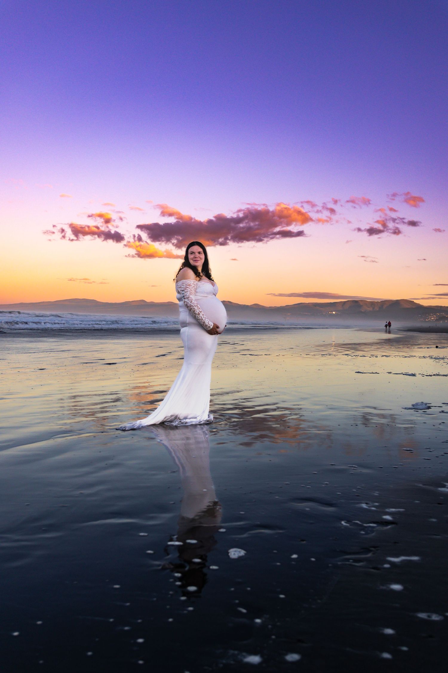 Pregnant woman in white lace gown standing on reflective beach at sunset with purple and orange sky.