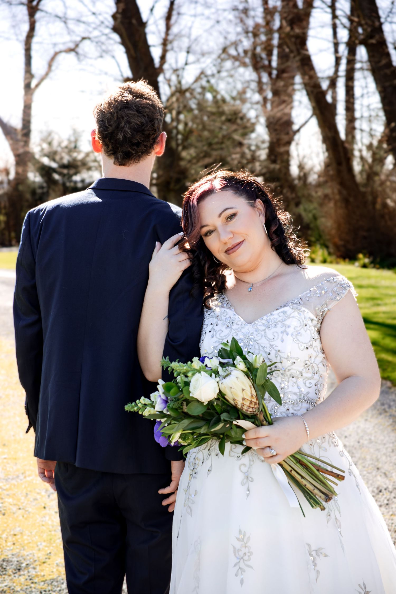Bride in white beaded gown holds floral bouquet beside groom in navy suit outdoors among trees.