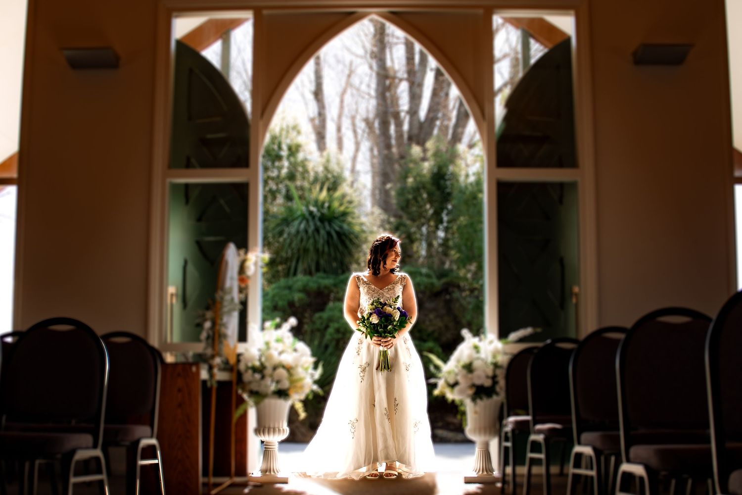 Bride in white gown stands in sunlit chapel doorway with floral arrangements and arched window backdrop.