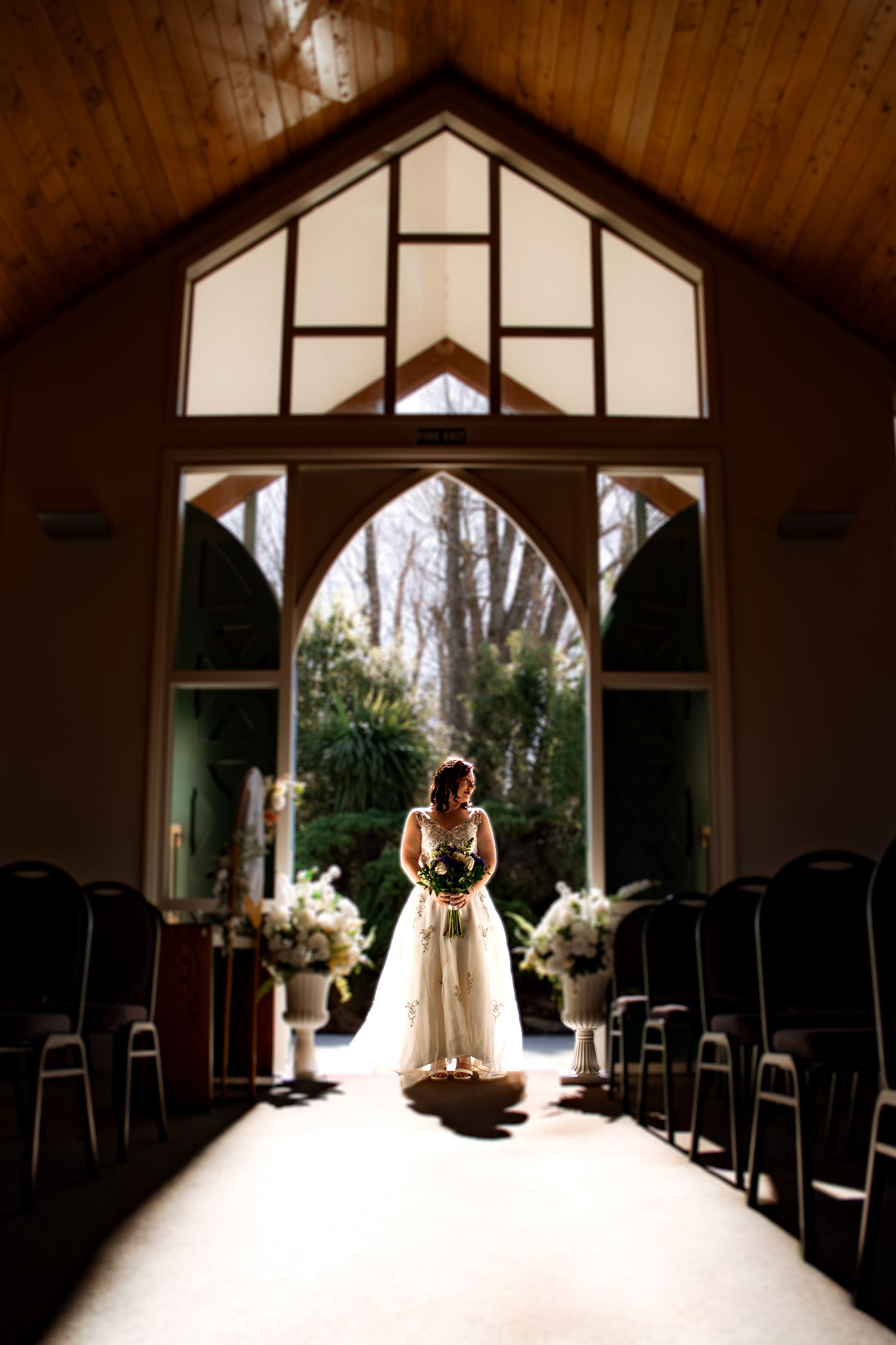 Bride in white gown stands at chapel altar, backlit by arched window, holding bouquet in elegant aisle setting.