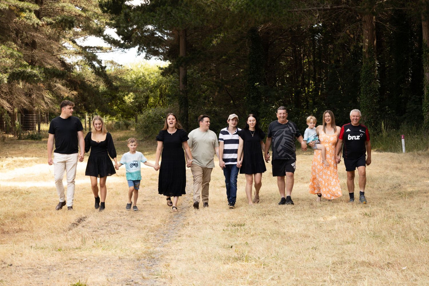 Large family group walking together on a sunny forest path surrounded by tall pine trees.