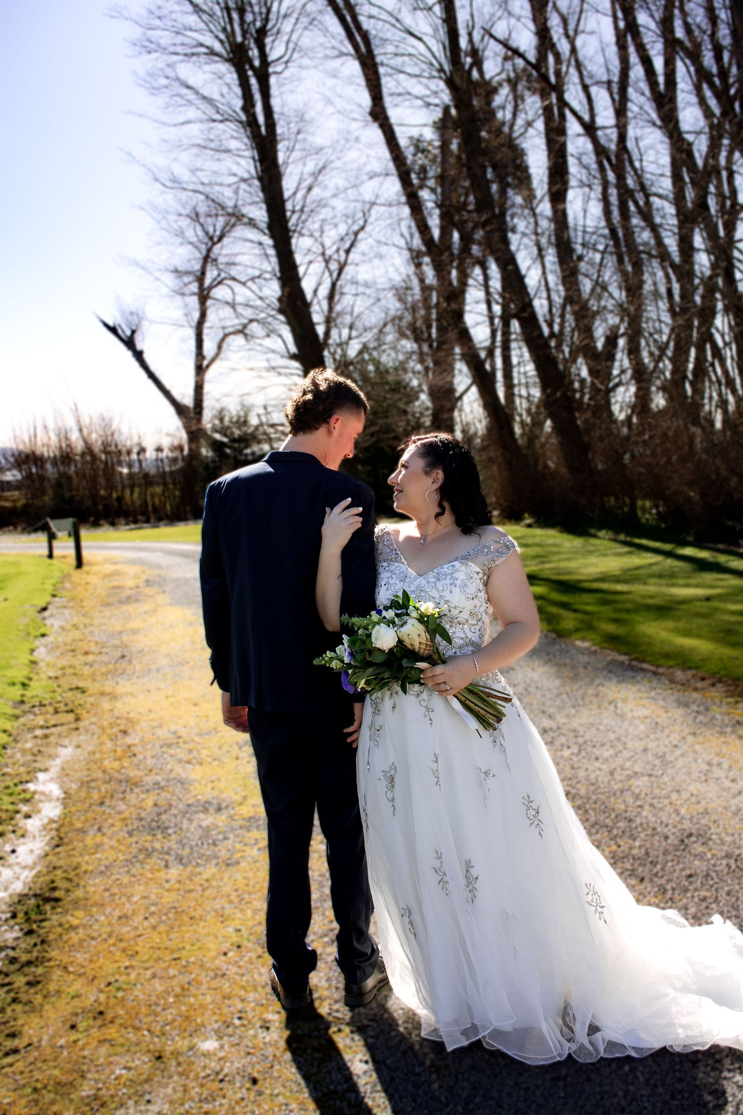 Bride and groom share a romantic moment on a gravel path surrounded by bare trees and green grass on their wedding day.
