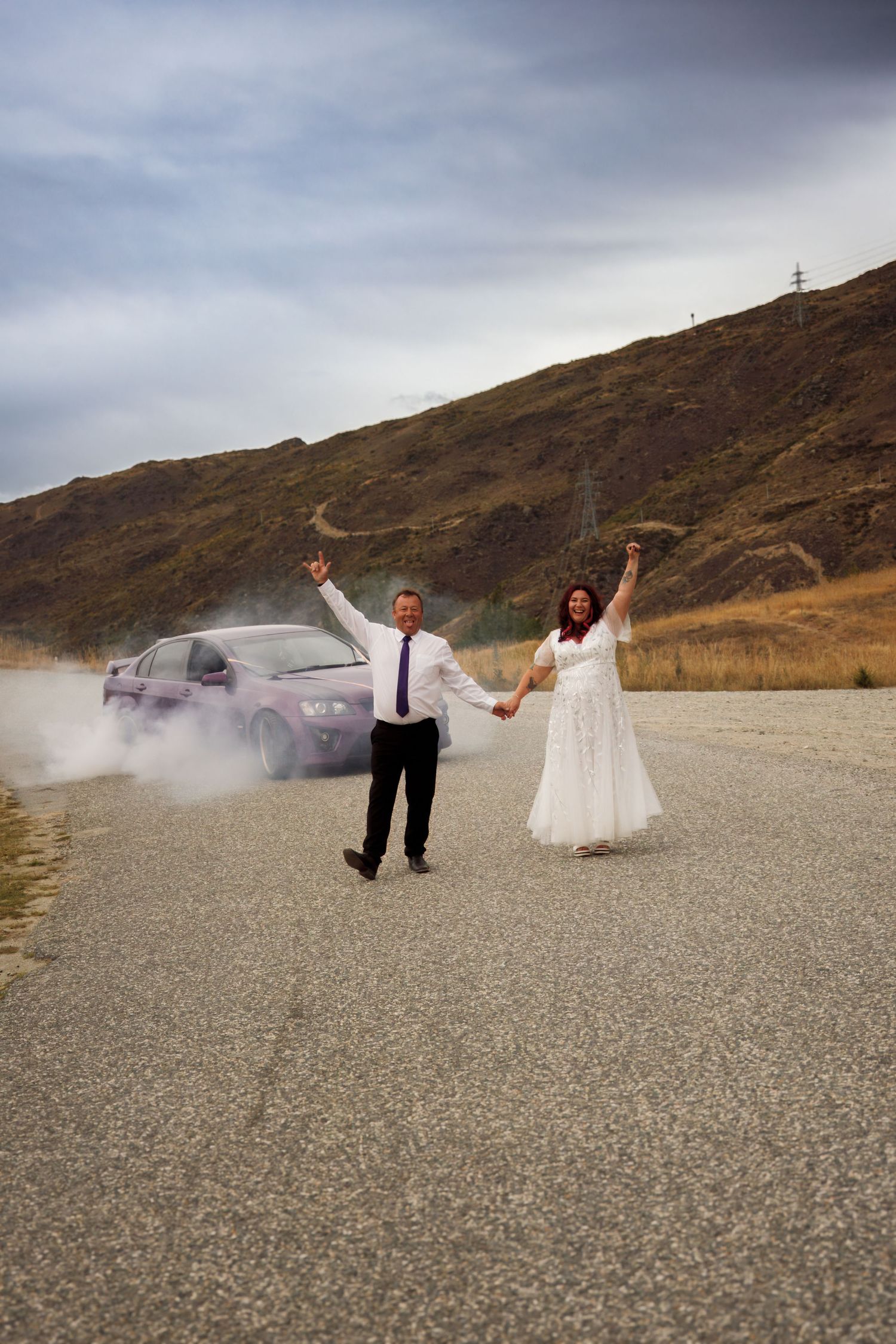 Bride and groom celebrate joyfully on open road with smoking purple car and mountain backdrop.