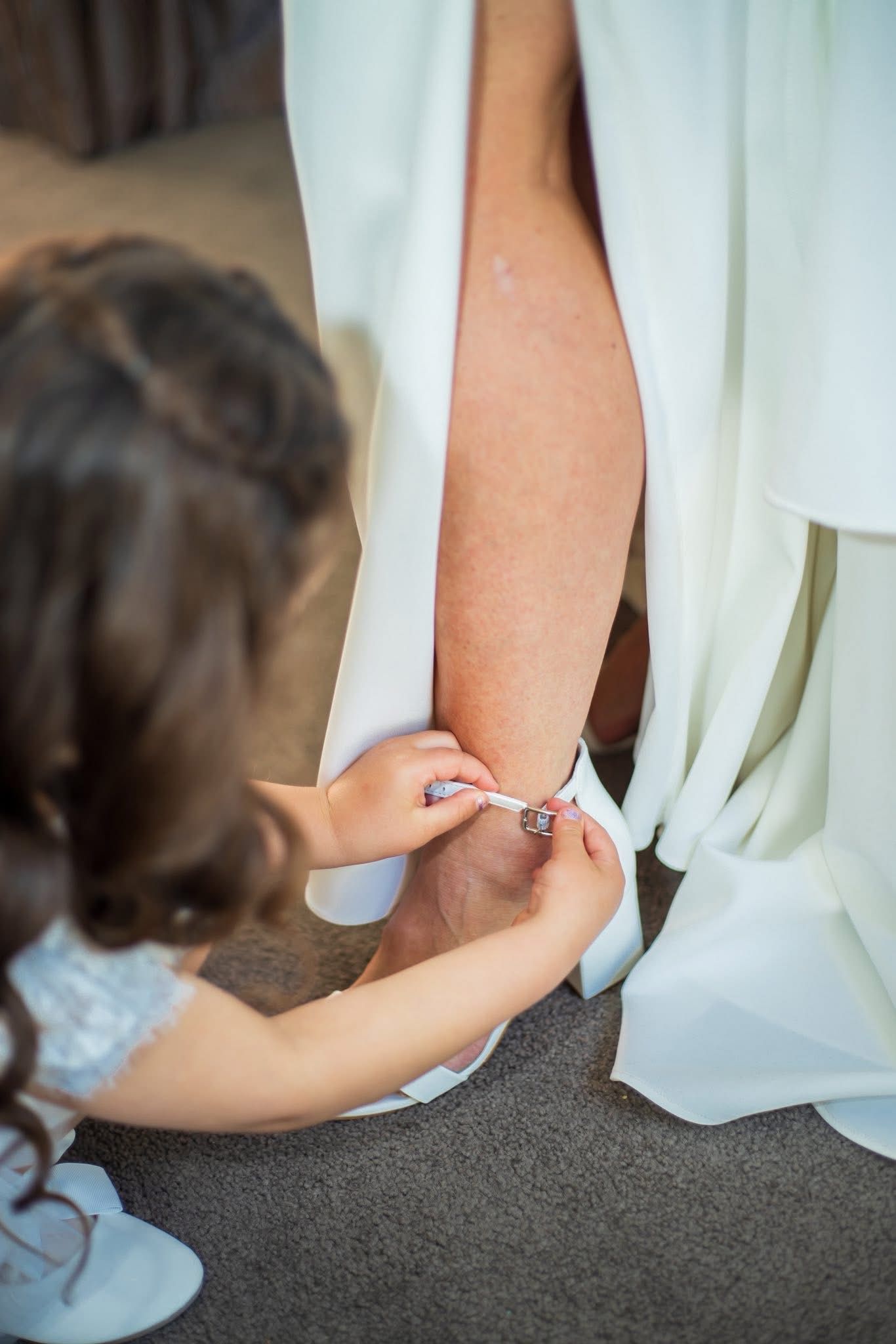 Young flower girl helping bride fasten her wedding shoe, a touching pre-ceremony moment captured beautifully.