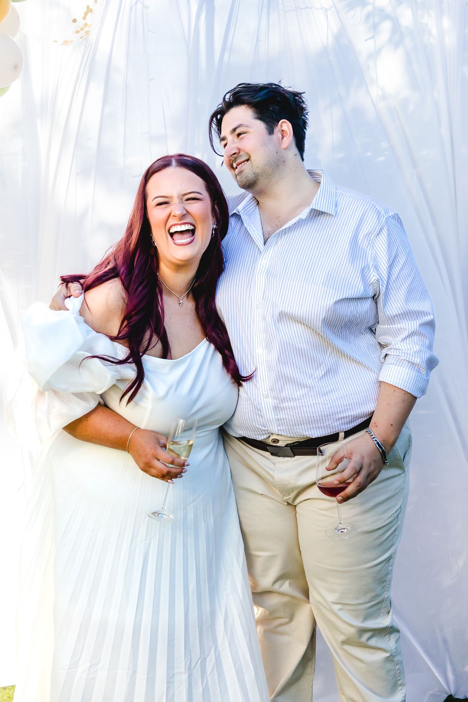 A joyful couple laughing together outdoors, woman in white dress holding champagne, man in light blue shirt and khaki pants.