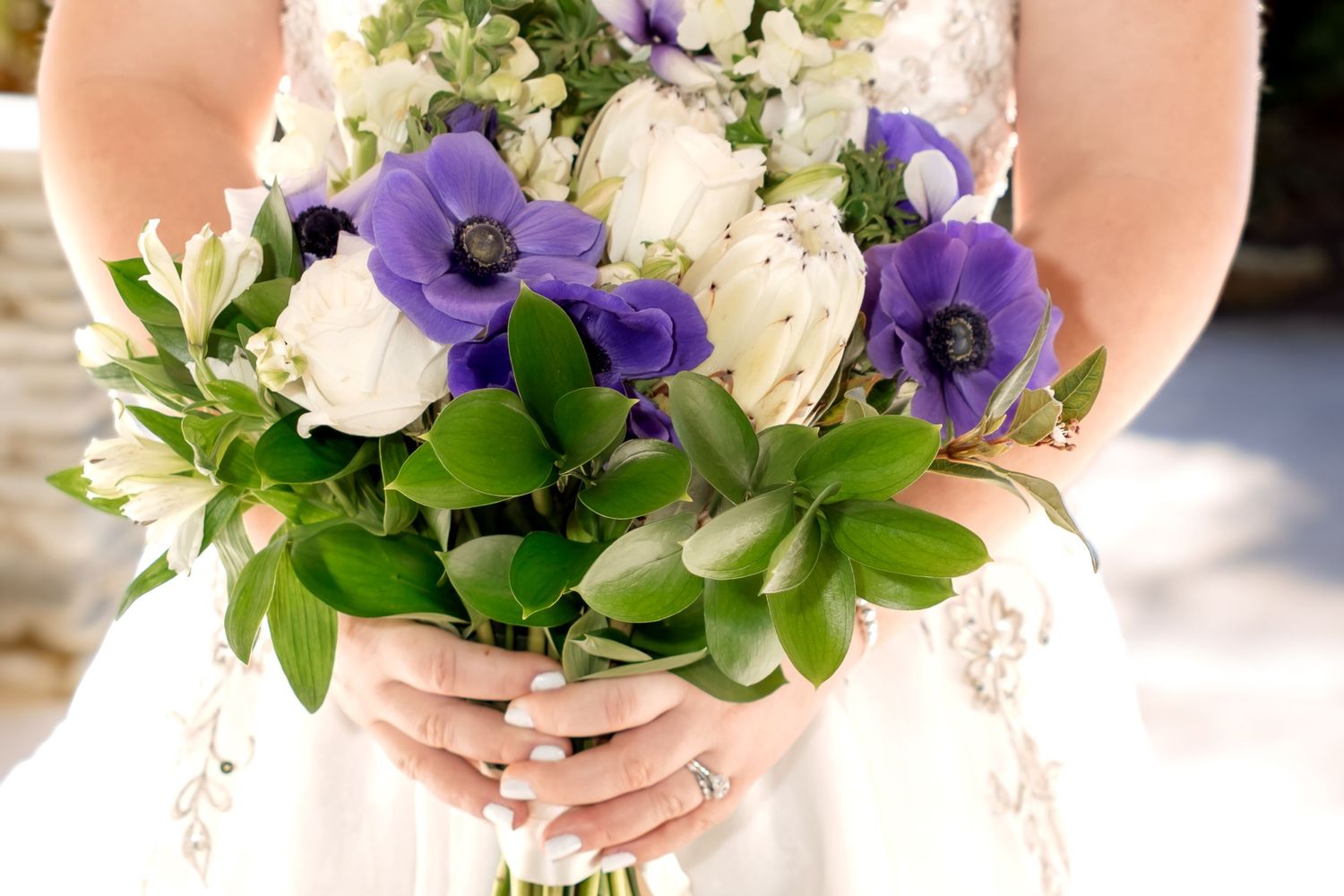 Bride holding a wedding bouquet with purple anemones, white tulips, and green succulents in a lush floral arrangement.