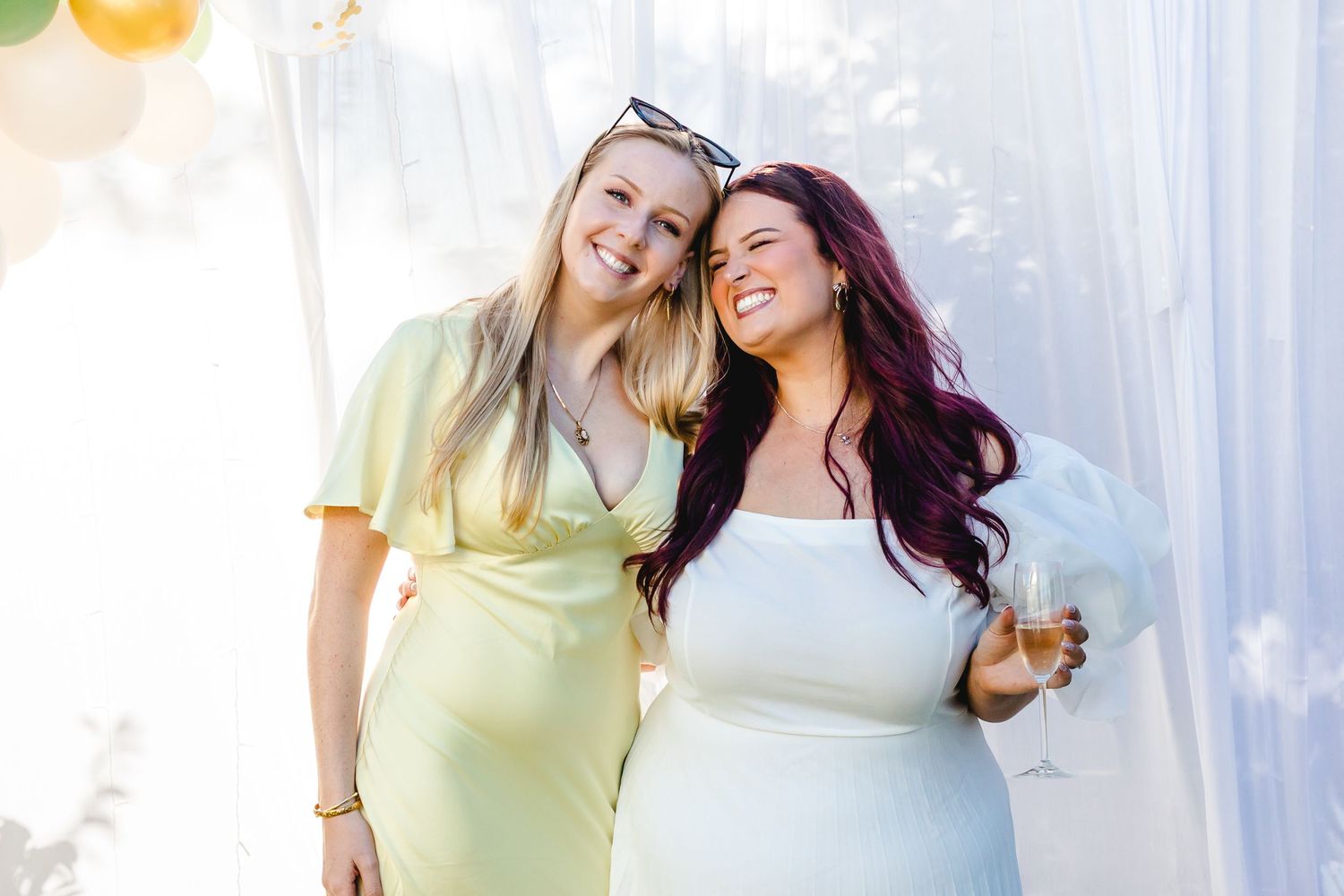 Two smiling women in elegant dresses pose together at a bright outdoor celebration, one holding a champagne glass.