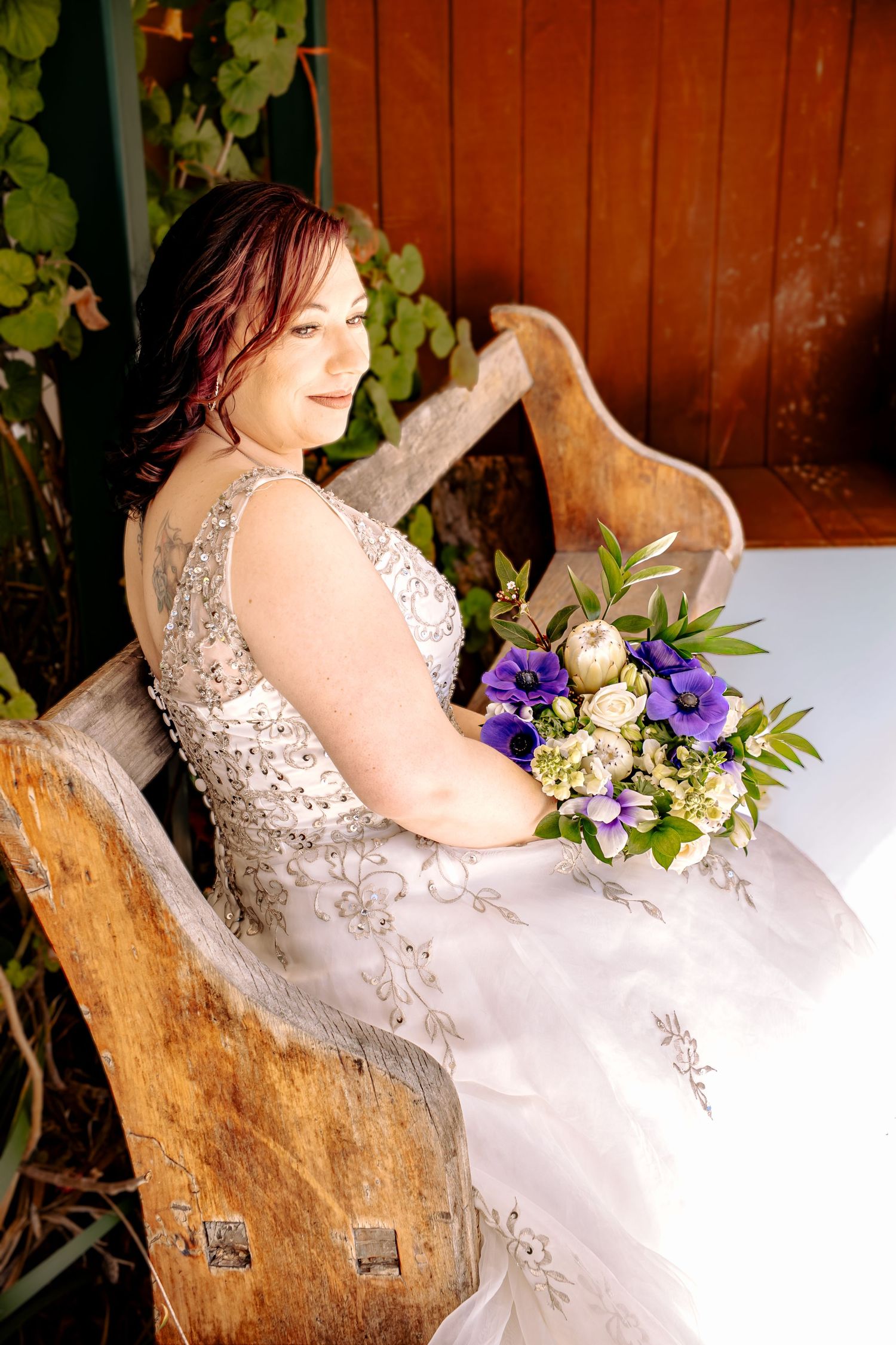 Bride in beaded gown sits on rustic wooden bench holding purple and white bouquet outdoors.