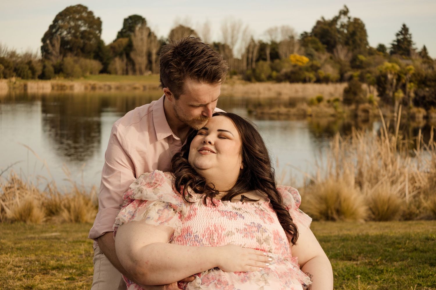Couple embracing outdoors by a serene lake, man kissing woman's forehead in a lush green park setting.