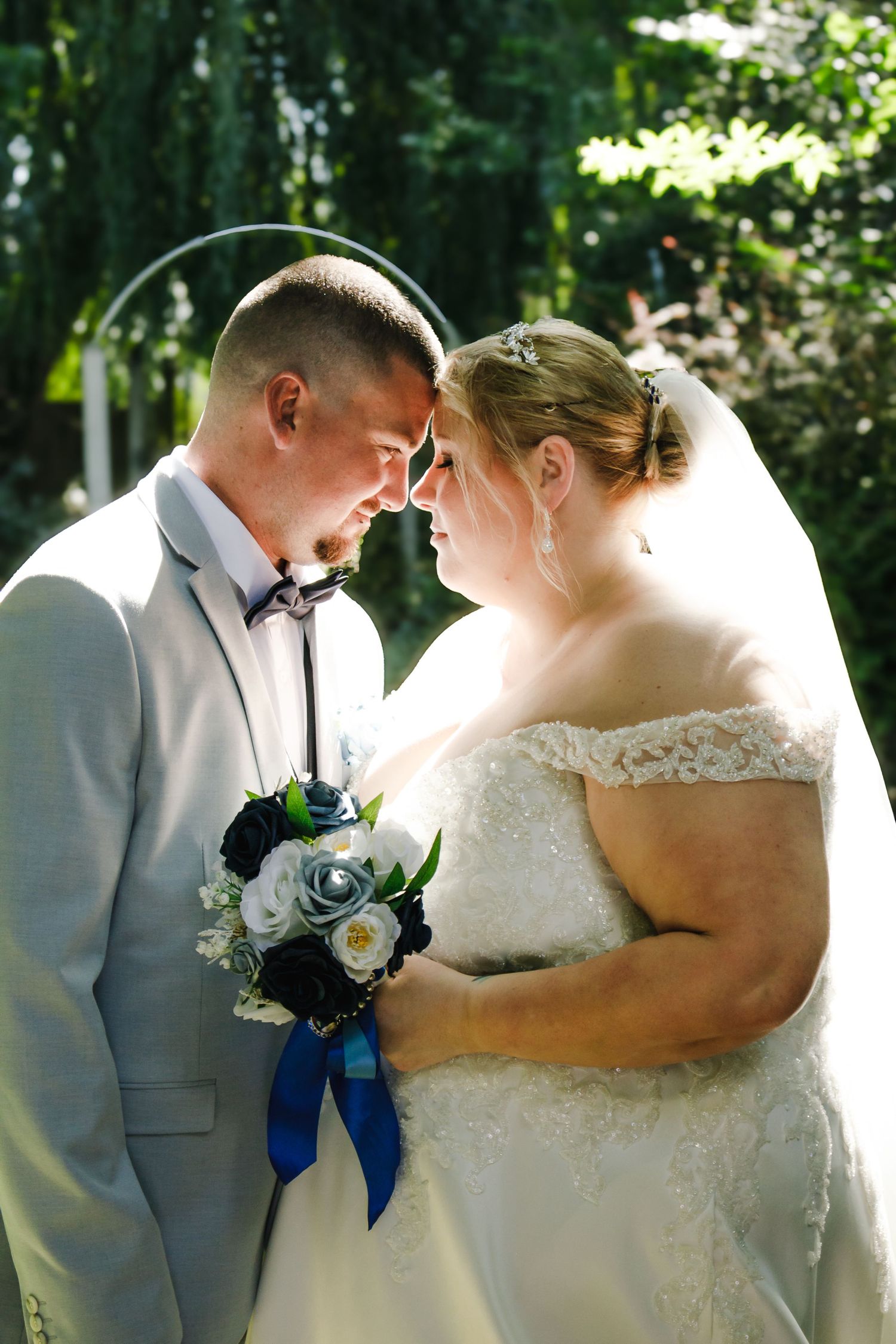 A tender moment between a couple in wedding attire standing close together in a sunlit garden setting.