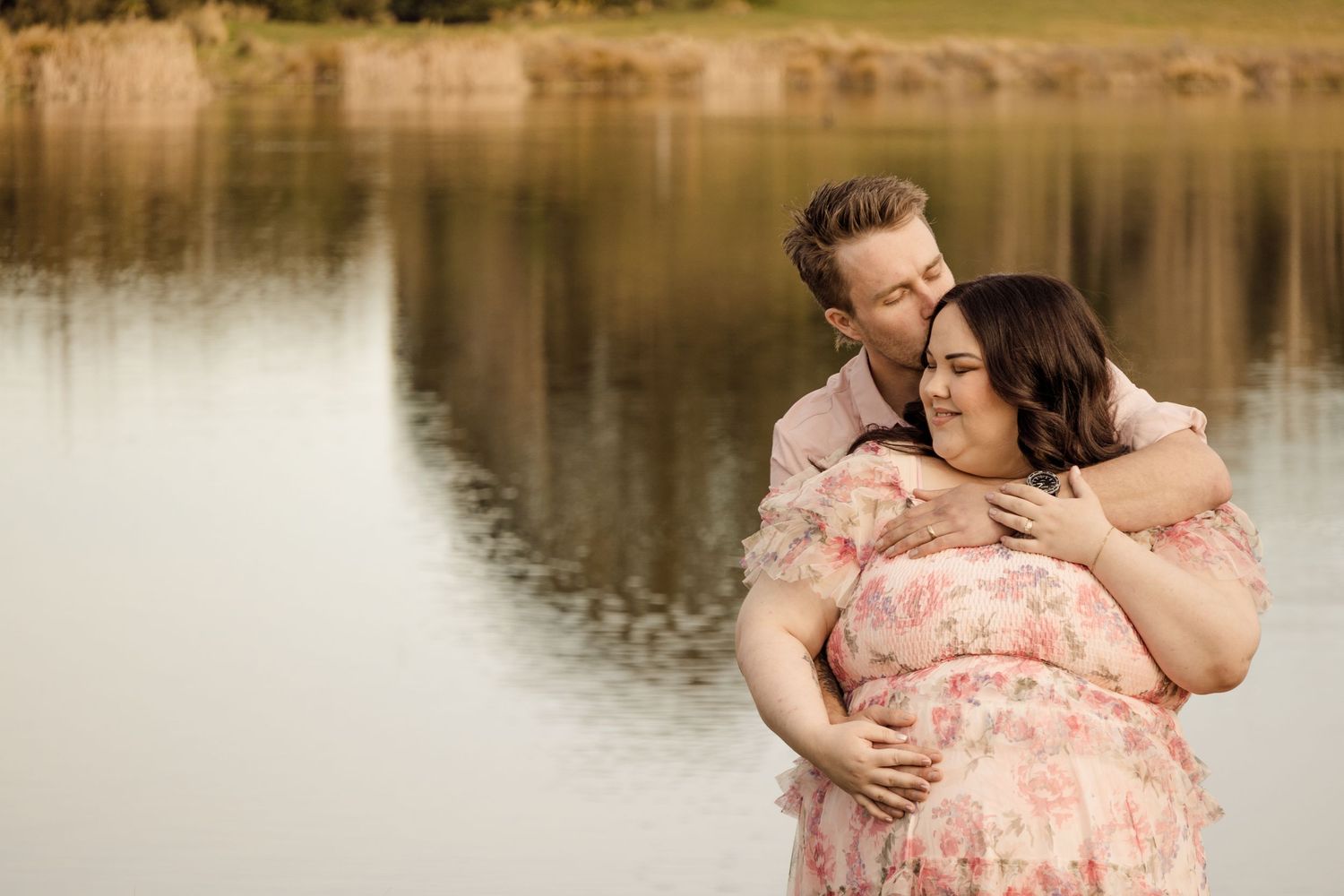 Couple embracing by a serene lake, pregnant woman in floral pink dress, man kissing her cheek outdoors.