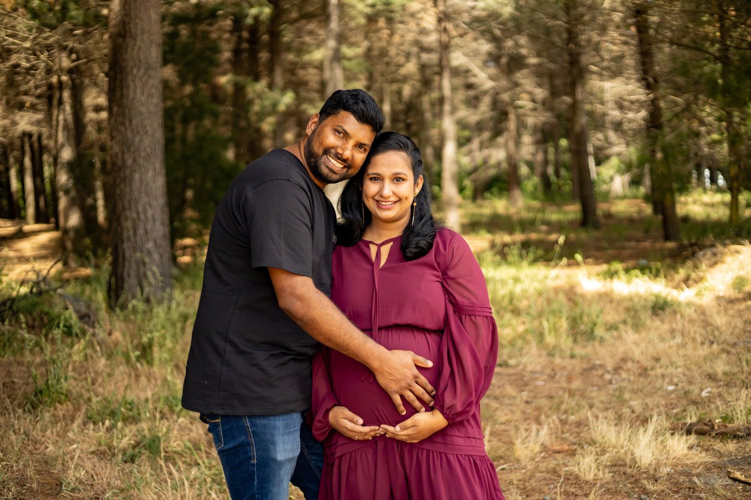 Couple posing outdoors in forest, pregnant woman in burgundy dress with partner in black shirt holding her baby bump.