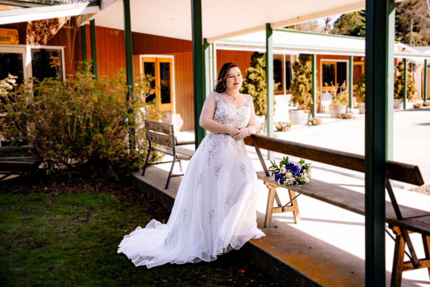 Elegant bride in a lace wedding gown posing outdoors at a rustic venue holding a blue bouquet.