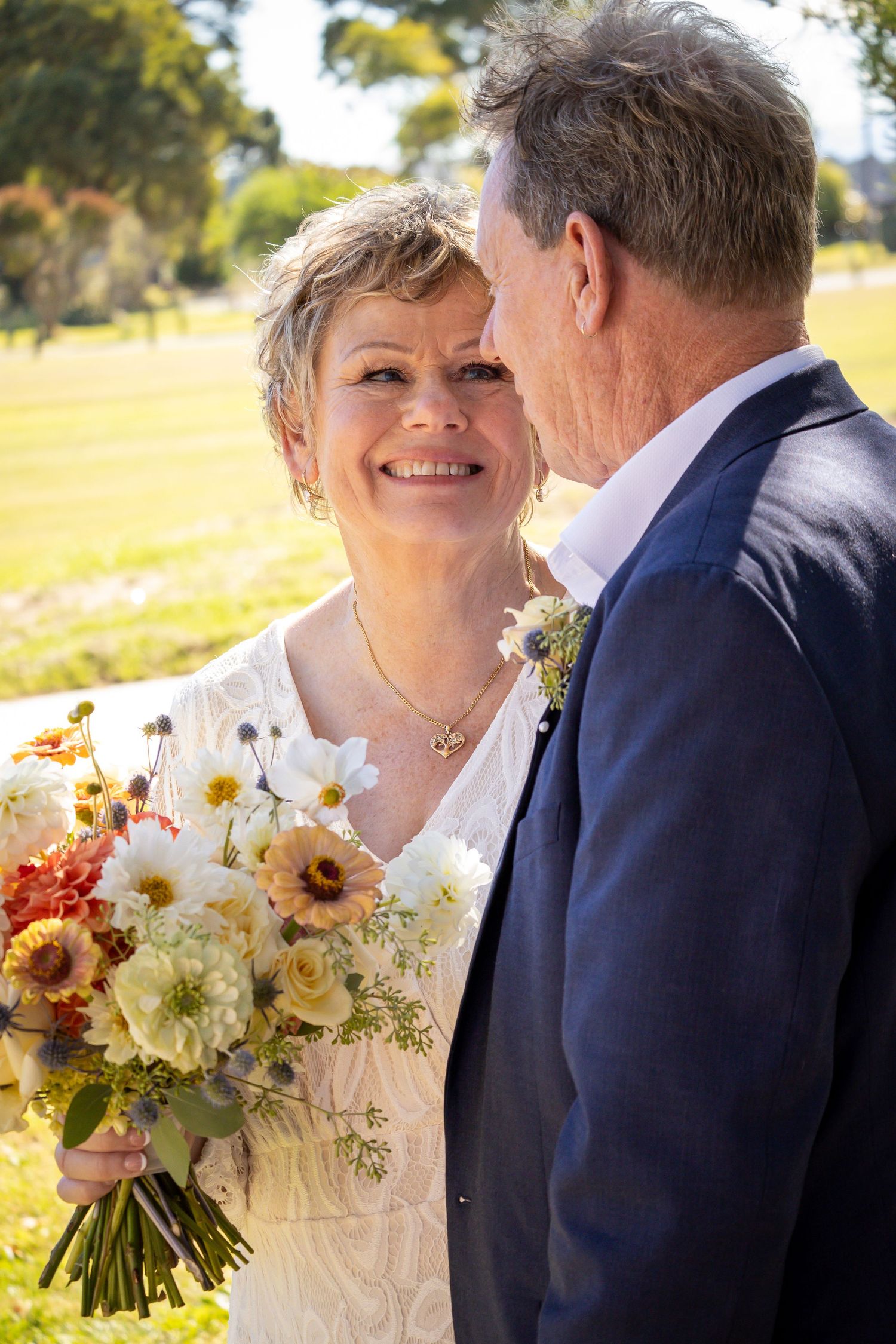 Smiling mature bride holding colorful bouquet stands close to groom in navy suit outdoors on sunny wedding day.