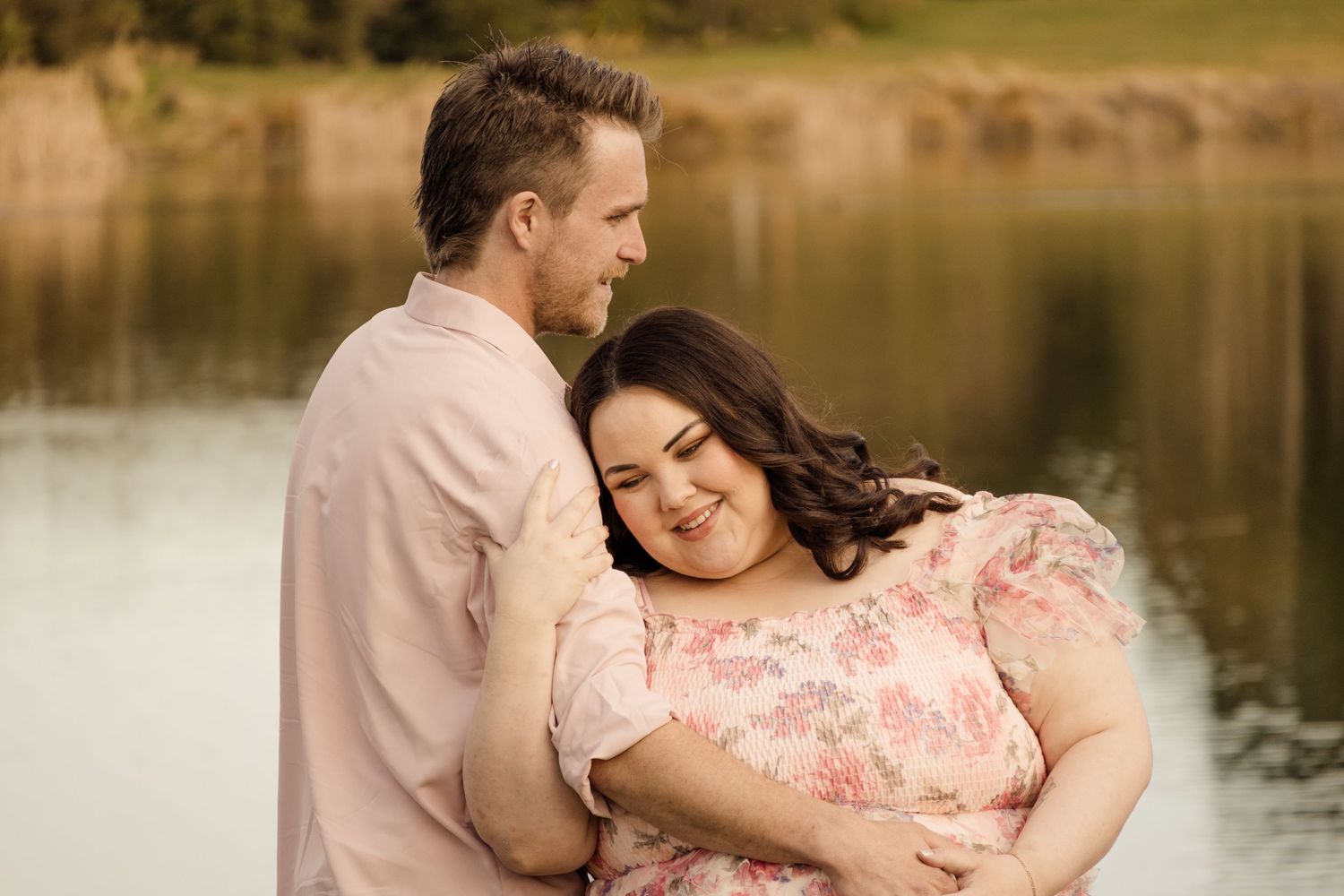 Couple embracing outdoors near a lake, woman in floral dress smiling, man in pink shirt, romantic engagement photo.