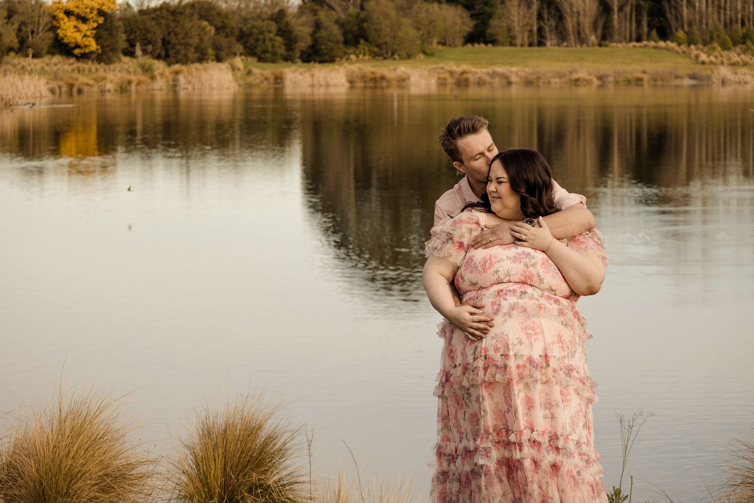Couple embracing by a serene lake, pregnant woman in floral pink dress, maternity photo shoot outdoors.