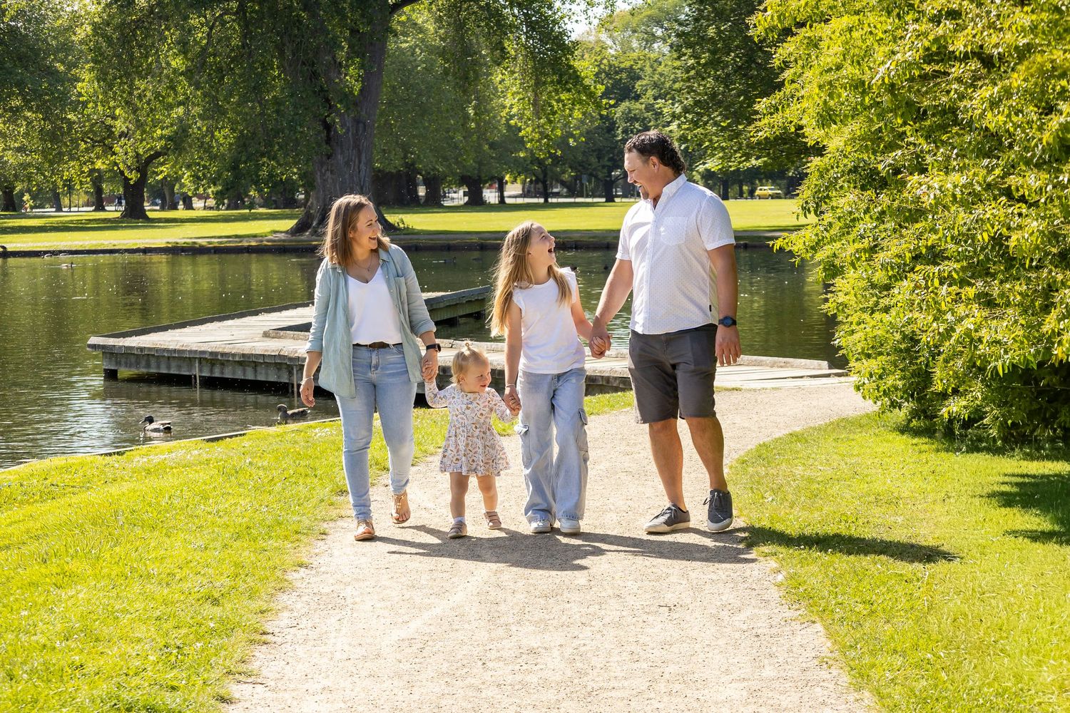 Family of four walking hand-in-hand along a sunny park path beside a calm pond on a bright summer day.