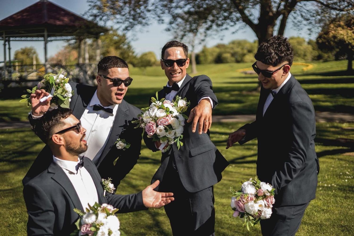 Four groomsmen in dark suits and sunglasses laughing and holding floral bouquets outdoors at a wedding.