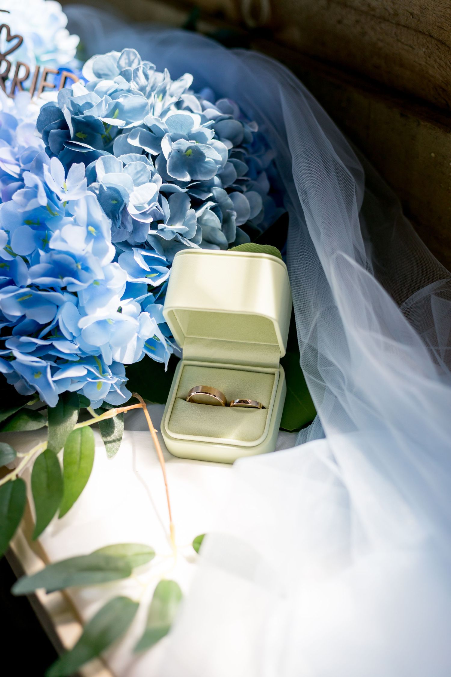 Gold wedding rings in a velvet box beside blue hydrangea flowers and a bridal veil on a wedding day.